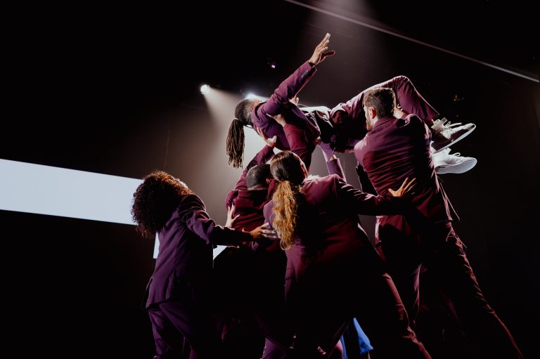Dance ensemble reaching upwards in dark red costumes